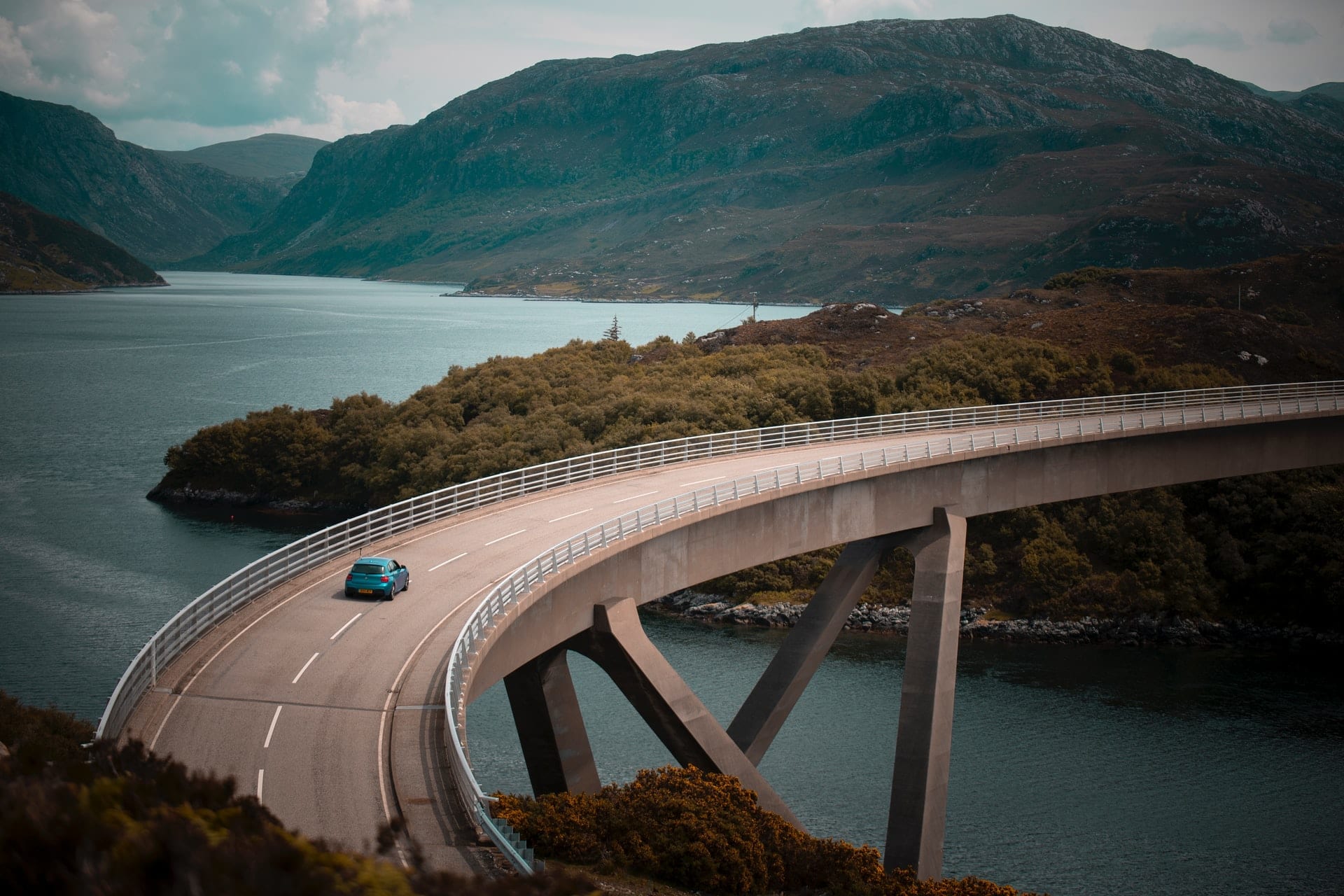 car-driving-across-a-winding-bridge-above-lake-with-mountains-in-background-kylesku-bridge-in-assynt-north-coast-500-scotland-best-scenic-drives-in-uk