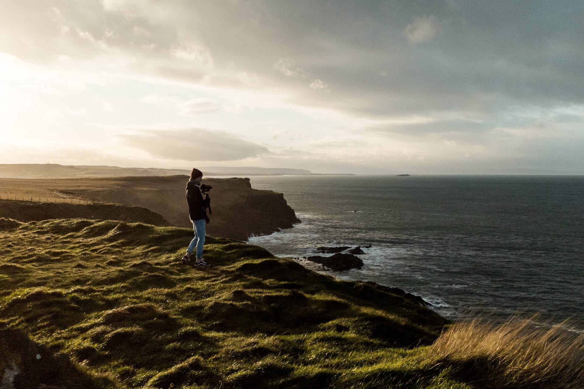 man-holding-camera-walking-along-cliff-by-coast-at-sunset-coastal-causeway-route-northern-ireland