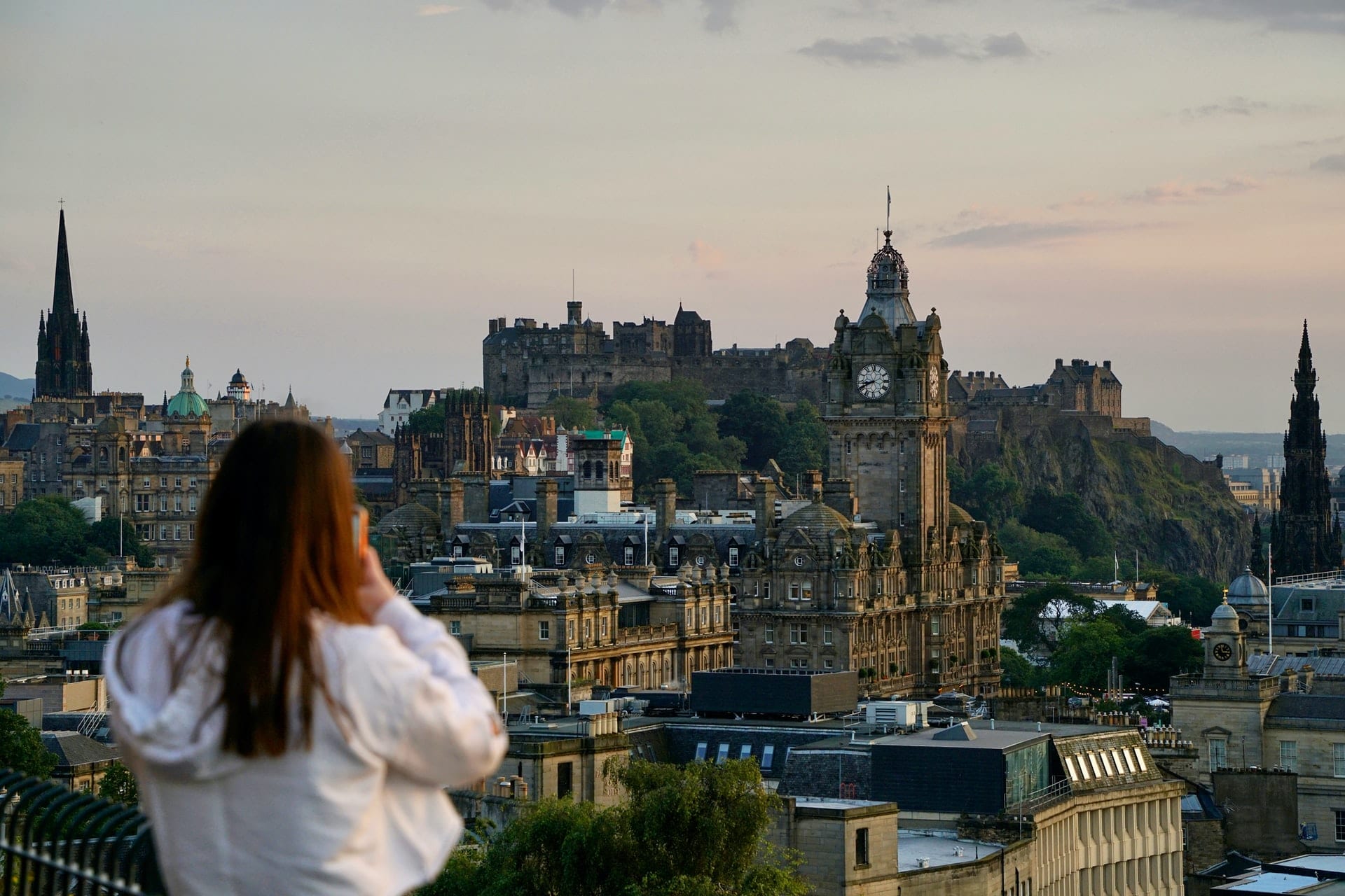 young-woman-standing-on-top-of-calton-hill-taking-a-photo-with-her-phone-of-edinburgh-city-edinburgh-castle-and-princes-street-gardens